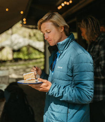 A woman wearing a blue puffer jacket with a visible 'dyrobe' logo, holding a slice of cake on a plate while standing under warm string lights.