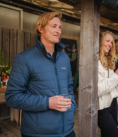 A man wearing a blue Dryrobe puffer jacket stands on a wooden porch holding a glass, with a woman in a white sweater partially visible behind a wooden post.