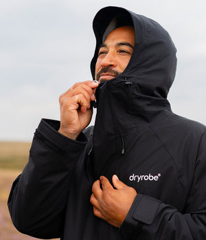 A man wearing a black hooded dryrobe jacket, adjusting the hood and zipper while standing outdoors against a cloudy sky.