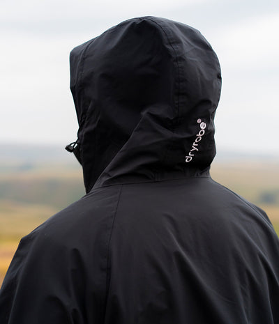*MALE* A person viewed from behind wearing a black hooded waterproof jacket with a small white Dryrobe logo on the side, set against a blurred outdoor landscape under an overcast sky.