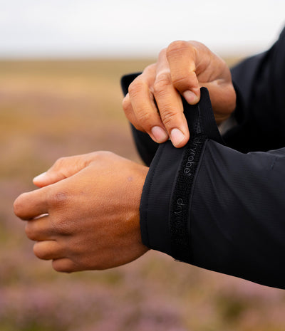 *MALE* Close-up of a person's hands adjusting the elasticated cuff of a black sleeve, with a blurred outdoor landscape in the background.