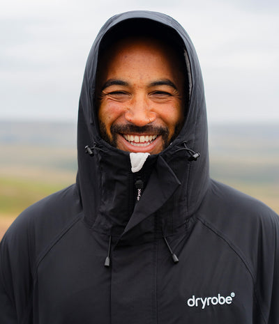 *MALE* A smiling man wearing a black Dryrobe hooded jacket with the brand logo on the left chest, set against a blurred outdoor landscape background.