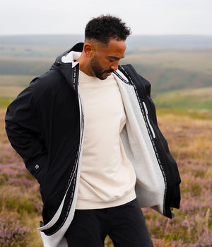 A man wearing a black hooded jacket with a white sherpa lining stands outdoors in a grassy, heather-covered landscape. The jacket features a black outer shell with white text detailing along the zipper edges and a white sherpa interior.