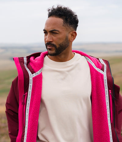 *MALE* A man wearing a bright pink fleece-lined jacket over a cream t-shirt, standing outdoors in a grassy field under an overcast sky.
