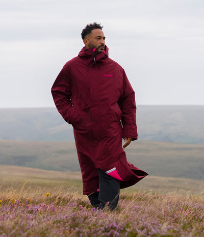 *MALE* A man wearing a long maroon waterproof parka and black trousers stands in a grassy, flowered landscape under an overcast sky.