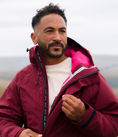 *MALE* A man wearing a maroon hooded technical jacket with a bright pink fleece lining, zipped open over a white t-shirt, set against a soft-focus outdoor landscape.