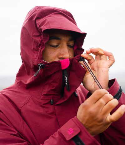 *MALE* A close-up of a man wearing a maroon hooded waterproof jacket, adjusting the drawstring of his hood with both hands in an outdoor setting.