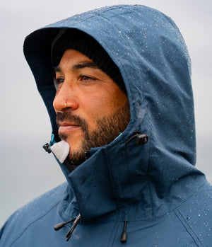 A close-up of a man wearing a dark blue hooded rain jacket with raindrops on it, set against a soft, overcast gray background.