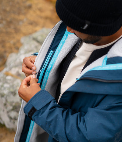 *MALE* A close-up lifestyle shot of a man wearing a blue and gray fleece-lined jacket, adjusting the zipper on the chest. The jacket features a bright blue zipper track with white text that reads 'dryrobe'.