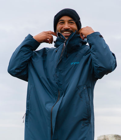 *MALE* A man wearing a blue Drybot waterproof jacket and a dark knit beanie, smiling and adjusting the collar of the jacket against an overcast sky.
