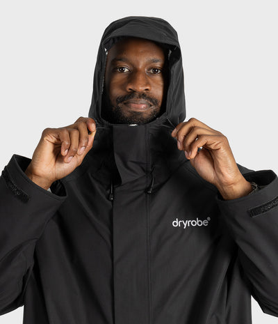 *MALE* A man wearing a black hooded Dryrobe jacket, holding the collar of the hood with both hands against a light gray studio background.