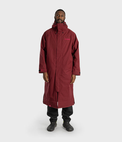 *MALE* A man wearing a long maroon hooded technical parka with a small pink logo on the left chest, paired with black pants and dark shoes, standing against a light gray studio background.