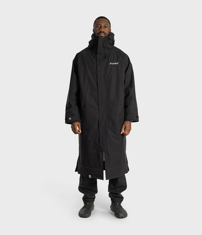 *MALE* A man wearing a long black hooded coat with a white 'branded' logo on the left chest, black pants, and black shoes, standing against a light gray studio background.