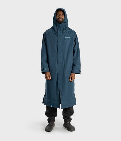 *MALE* A man wearing a long, hooded waterproof coat in a muted blue-gray color, paired with black pants and boots, standing against a light gray studio background. The brand name 'Dryrobe' is visible on the left chest of the coat.