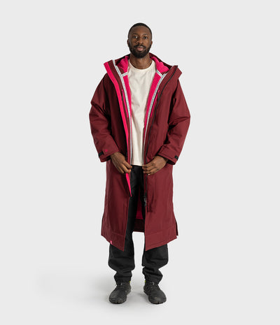 *MALE* A man wearing a long burgundy hooded parka with a bright pink inner lining and zipper trim, layered over a white t-shirt, black pants, and black boots, standing against a light gray studio background.