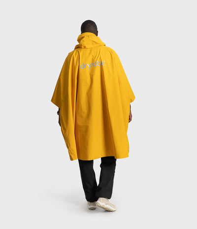 *MALE* A model viewed from behind wearing a bright yellow hooded poncho with the brand name 'dryrobe' printed in white on the back, paired with black pants and white sneakers, against a light gray studio background.