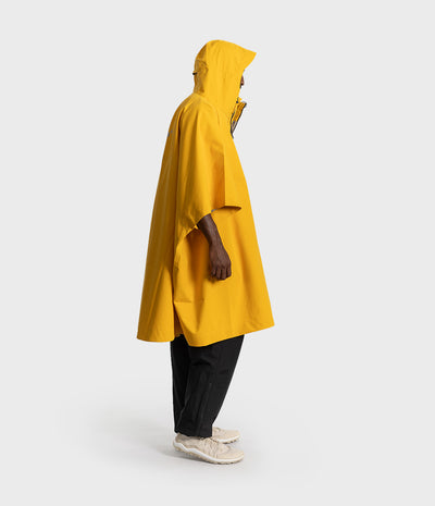 *MALE* A person shown in side profile wearing a bright yellow hooded rain poncho over black pants and light-colored sneakers, photographed against a clean light gray studio background.