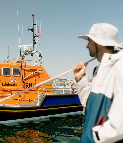 *MALE* A man in a white bucket hat and grey Dryrobe stands in the foreground holding a rope, with an orange lifeboat and blue water in the background under a clear sky.