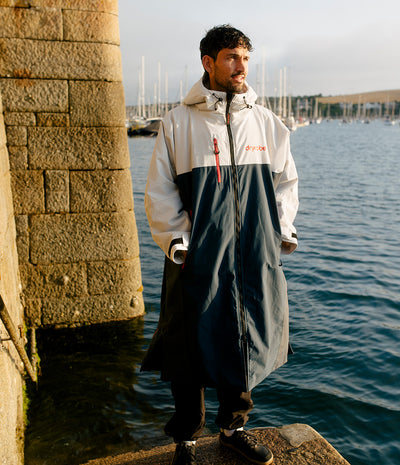 *MALE* A man wearing a long, two-tone waterproof Dryrobe stands on a stone pier next to the water, with a marina in the background. The raincoat is white and dark grey with a red logo on the chest.