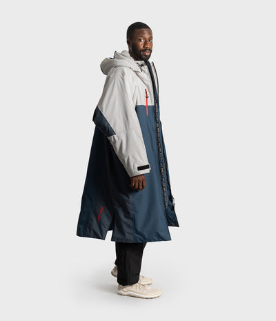 *MALE* A man wearing a long hooded colour-blocked Dryrobe in light grey and navy blue, paired with black pants and off-white sneakers, standing against a light grey studio background.