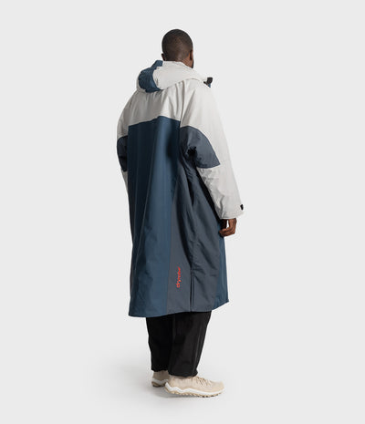 *MALE* A man viewed from behind wearing a long hooded Dryrobe in white and navy blue, paired with black pants and beige sneakers, against a light grey studio background.