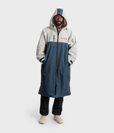 *MALE* A man wearing a long hooded raincoat in light grey and navy blue with a red zipper and red 'Dryrobe' logo on the chest, paired with black pants and beige sneakers, against a light grey studio background.