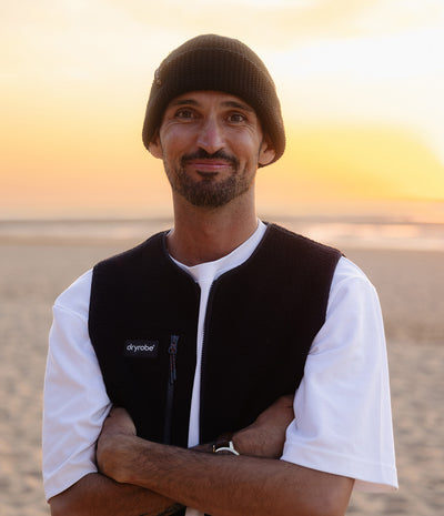 *MALE* A man wearing a black knit beanie, a black Dryrobe fleece vest over a white t-shirt, standing on a sandy beach at sunset.