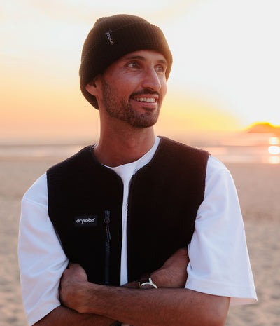 *MALE* A man wearing a black knit beanie and a black Dryrobe fleece vest over a white t-shirt, standing on a beach at sunset.