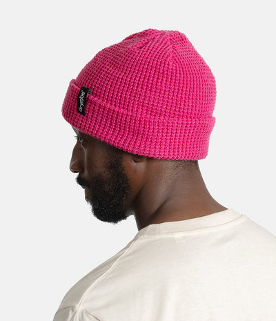 A man in profile wearing a bright pink ribbed knit beanie with a small black brand patch on the cuff, photographed against a clean light gray background.