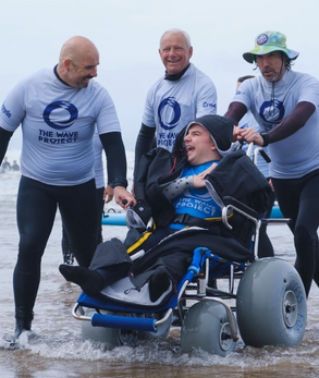 Man in wheelchair in ocean with 3 men standing behind him laughing