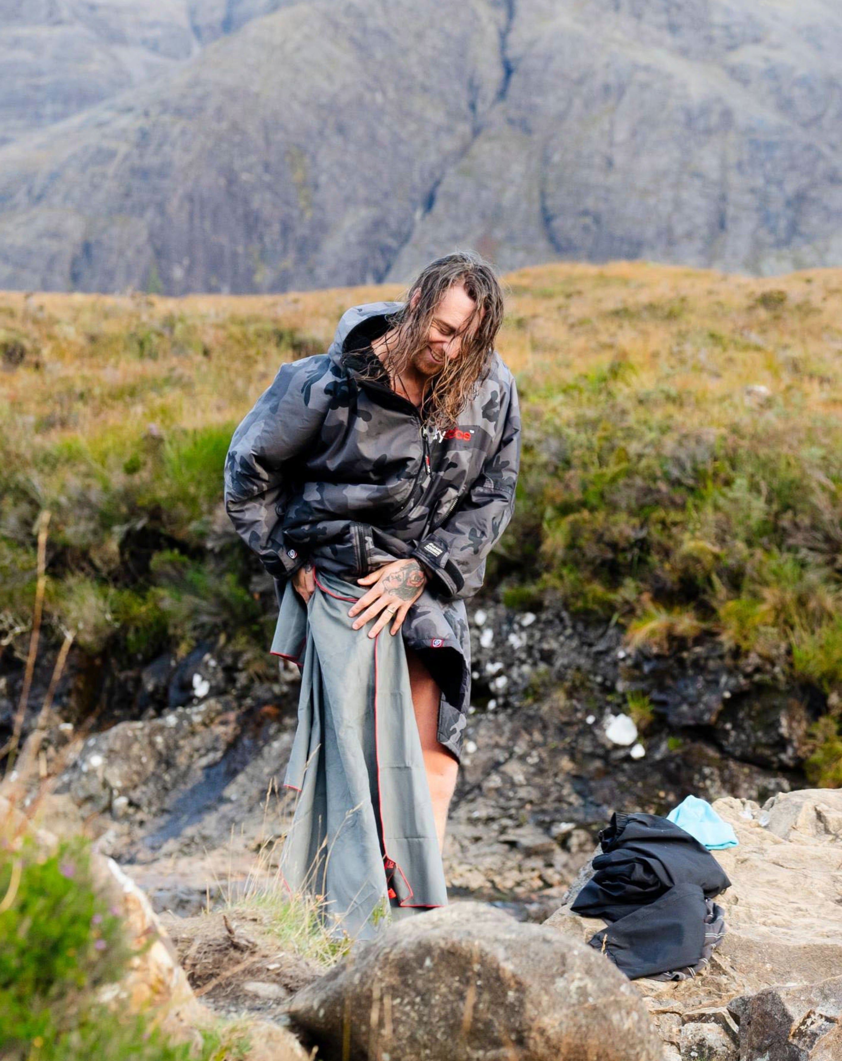 A man in a wet, dark camouflage Dryrobe stands on a rocky, grassy mountainside.