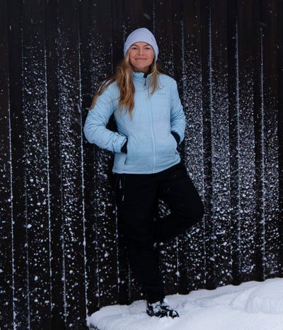 A woman wearing a light blue ski jacket, black ski pants, a white beanie, and black gloves stands in the snow against a dark wooden fence with falling snow.