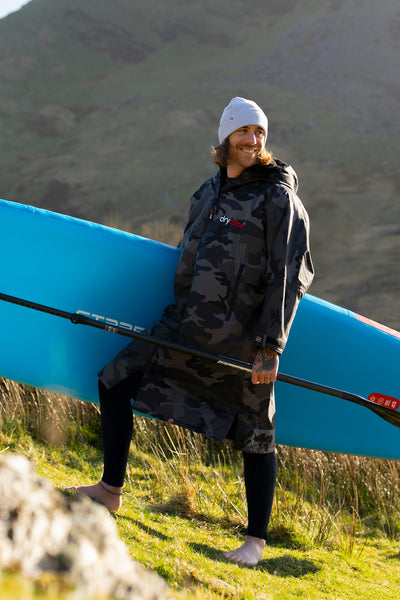 Person holding a blue paddleboard in a grassy field with mountains in the background