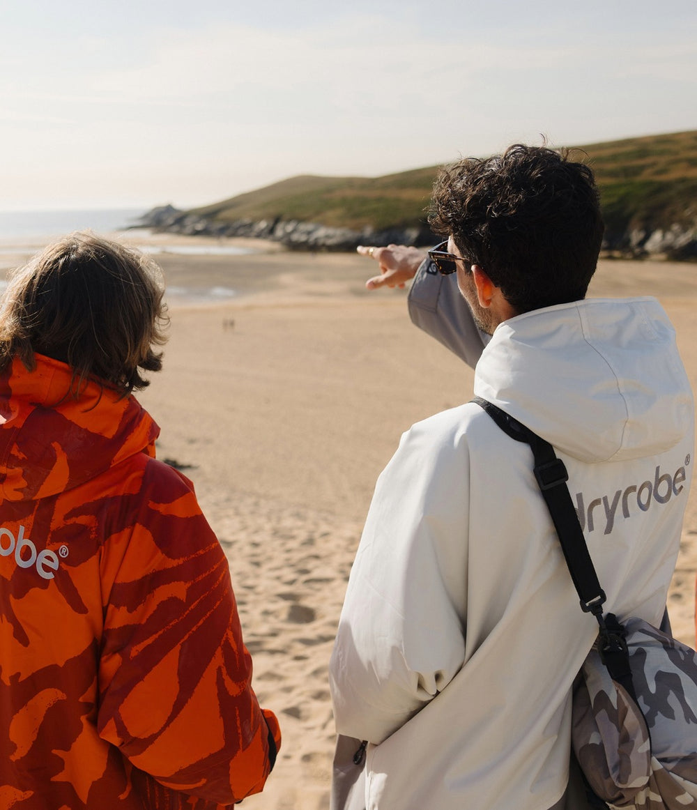 Two people standing on a sandy beach looking out toward the ocean and rocky coastline. The person on the right wears a white Dryrobe hoodie with a black shoulder bag, and the person on the left wears a bright orange and red patterned top.