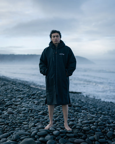 *MALE*Person wearing a black coat standing on a rocky beach with a cloudy sky.