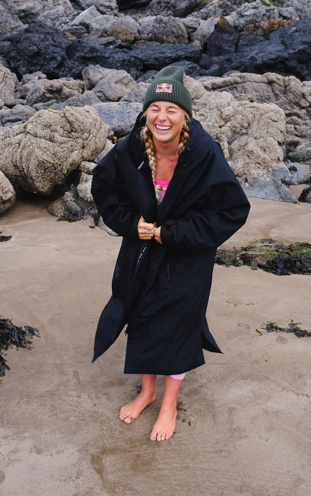 A woman standing on a sandy beach in front of large rocks, wearing a black Dryrobe Nexus over a pink swimsuit, with a green beanie.