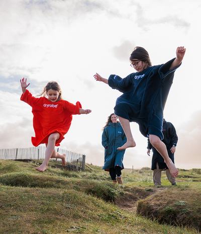 Two people in colourful towel robes jumping on a grassy hill with a cloudy sky.
