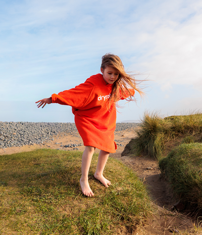 Child wearing an orange 'dryrobe' towel robe on a grassy path with a blue sky background