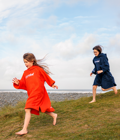 Two children running on a grassy area by the water, wearing branded ponchos.