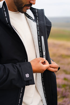 A close-up of a man wearing a black outdoor jacket with a white sherpa lining, zipping up the front in an outdoor landscape setting.