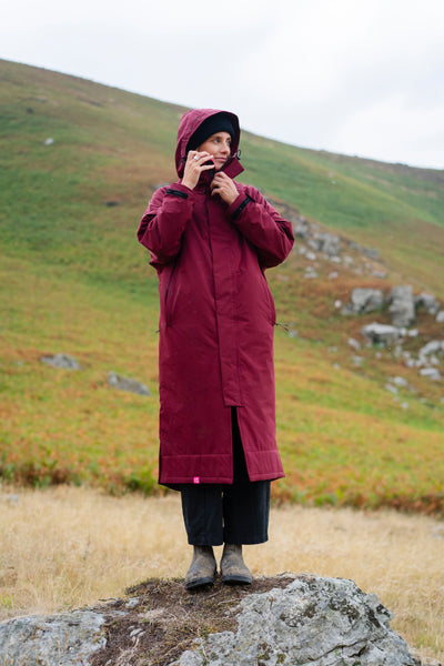 A person wearing a long burgundy hooded raincoat and dark pants stands on a large rock in an outdoor grassy landscape with hills in the background.
