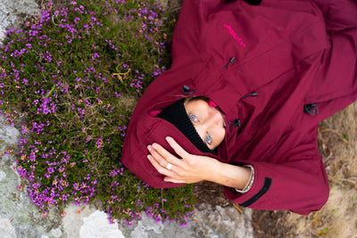 A person lying on their back outdoors on a rock, wearing a burgundy hooded jacket with a black beanie. The jacket has a pink 'KONTEK' logo on the chest.