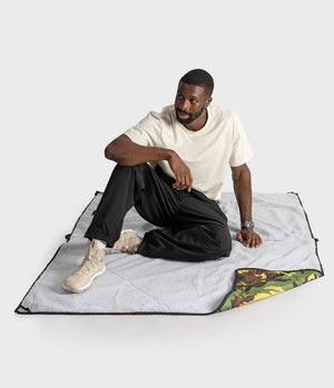 A man sits on a light grey picnic blanket with black trim, showing the reverse side with a green camouflage pattern, against a plain light grey studio background.