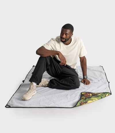 A man sits on a light grey picnic blanket with black trim, showing the reverse side with a green camouflage pattern, against a plain light grey studio background.