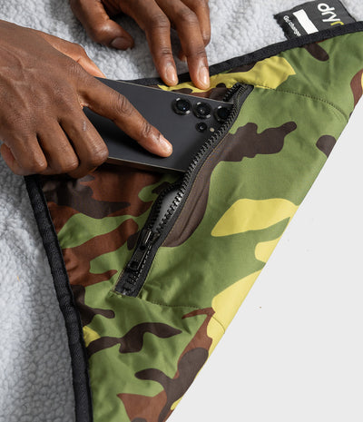 Close-up of a person's hands sliding a black smartphone into a zippered pocket of a camouflage fleece blanket. The blanket features a green, brown, and yellow camo pattern with black trim and a gray sherpa lining.