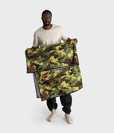 A man holding up a camouflage-patterned picnic blanket against a light grey studio background. The camouflage design features green, brown, black, and tan tones, with a black zipper running horizontally across the top.