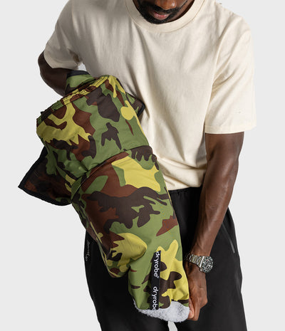 A person wearing a cream t-shirt and black pants holds a camouflage-patterned fleece-lined picnic blanket. The garment features a classic green, brown, and yellow camo pattern .
