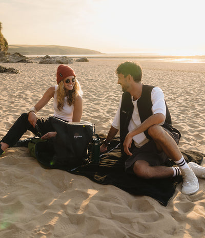 A man and a woman sit on a black backpack on a black blanket on a sandy beach at sunset. The woman wears a red beanie, white tank top, and black pants, while the man wears a white t-shirt, black vest, and white sneakers.