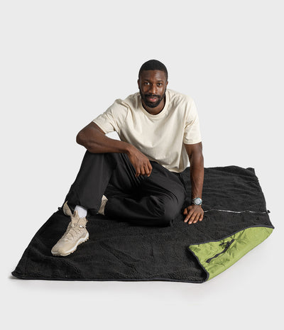A man sits cross-legged on a black textured blanket with a green underside, wearing a cream t-shirt, black pants, and beige sneakers, against a light grey studio background.