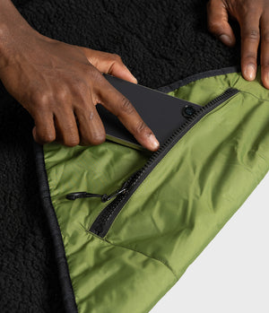 Close-up of a person's hands demonstrating a zippered pocket on an olive green picnic blanket, with a black fleece lining visible underneath.